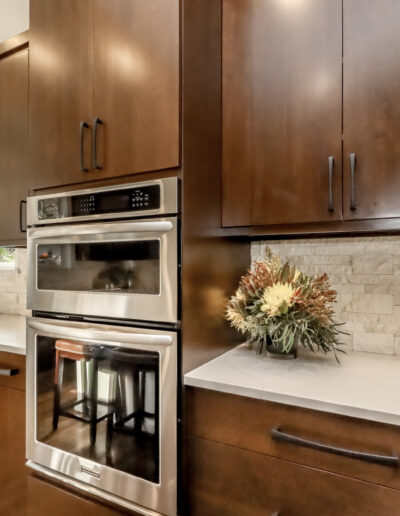 Modern kitchen with dark wood cabinets, white stone countertop, and a built-in stainless steel oven. A vase with flowers is on the counter near a window.