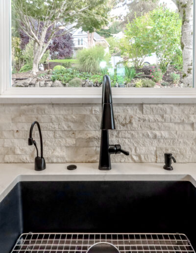 Black kitchen sink with modern faucets set in a white countertop. A large window above the sink shows a green garden view outside.
