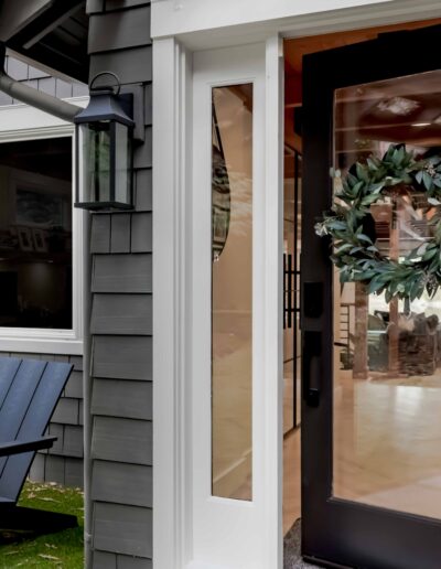 Black-framed front door with a green wreath, partially open, next to a window; two dark Adirondack chairs sit outside on grass.