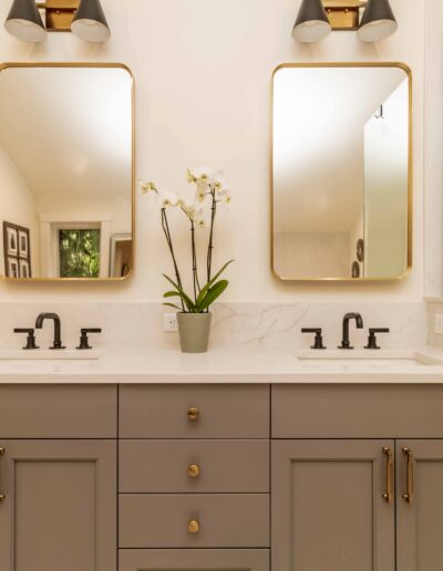 Modern bathroom with double sinks, gray cabinetry, gold hardware, two rectangular mirrors, black faucets, and a potted orchid on the countertop.
