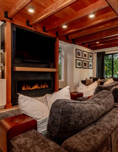Modern living room with exposed wooden beams, a built-in fireplace, sectional sofas, wall art, and large windows overlooking trees.