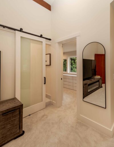 A modern bedroom with beige carpet, a sliding barn door, a tall arched wall mirror, and a view into an adjoining room with cabinets and a window.